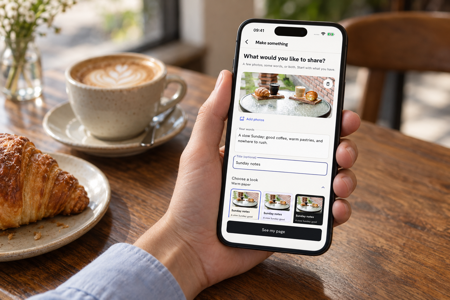 A hand holding the Edition app at a café, with a coffee and croissant on the table